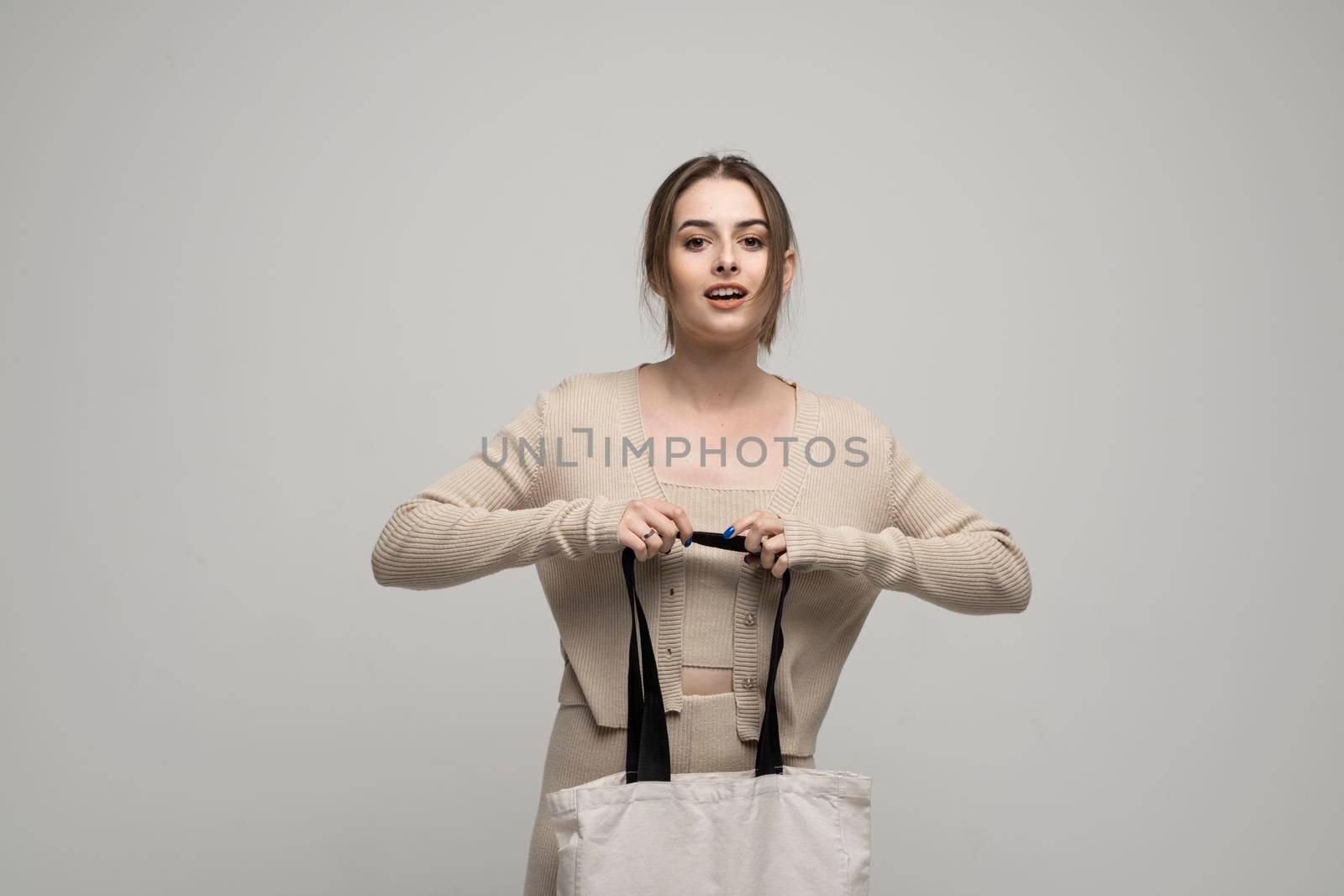 Beautiful brunette woman in a beige costume with cotton bag on in a hand on a grey background. Mockup and zero waste concept