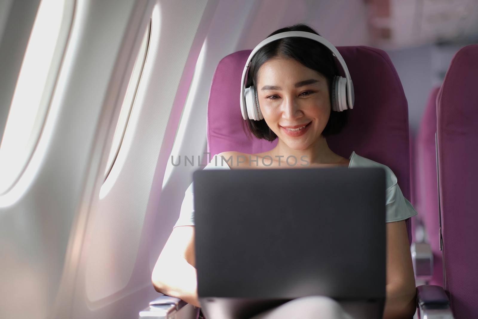 Asian young woman wearing headphone using laptop sitting near windows at first class on airplane during flight, Traveling and Business concept.