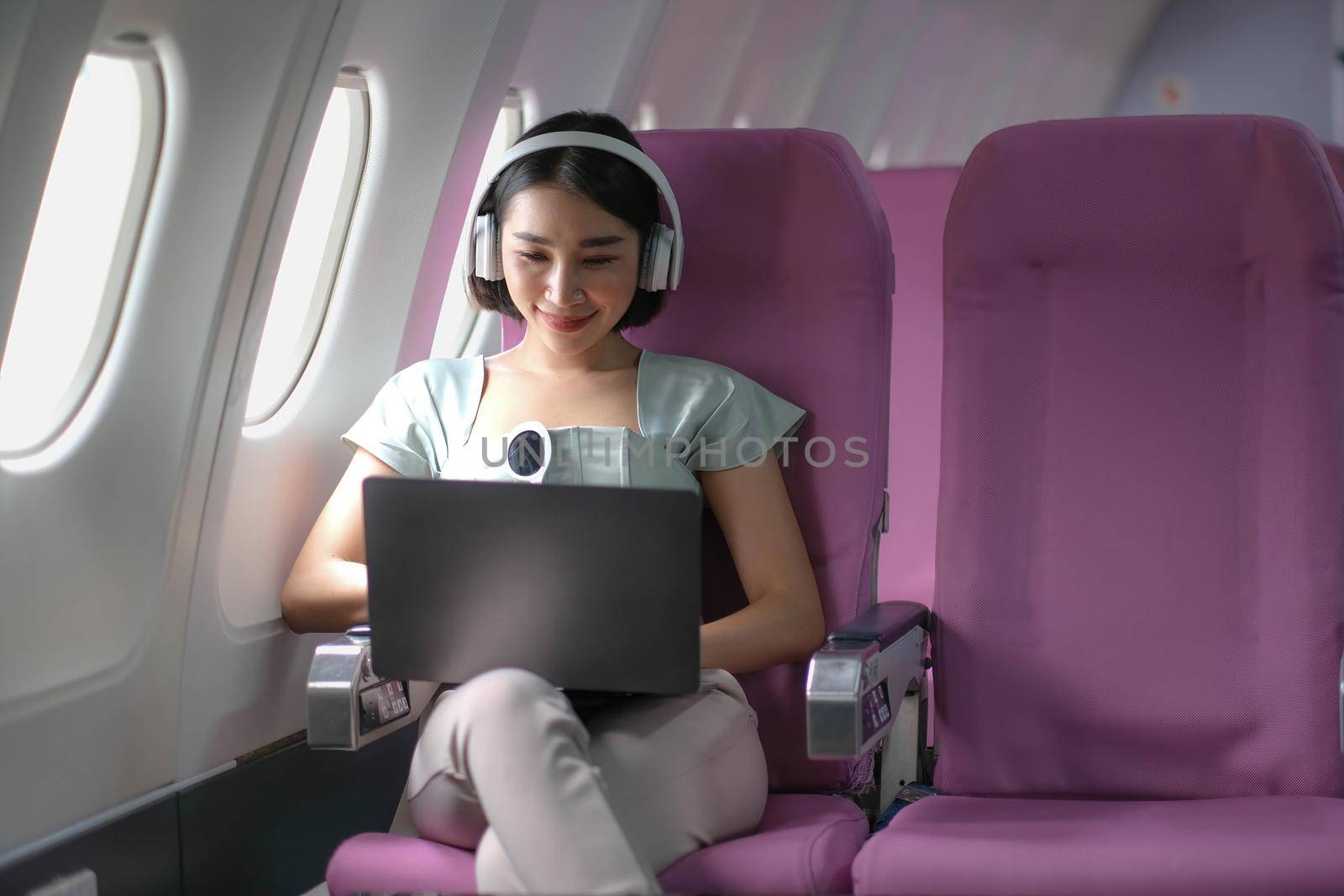 Asian young woman wearing headphone using laptop sitting near windows at first class on airplane during flight, Traveling and Business concept.