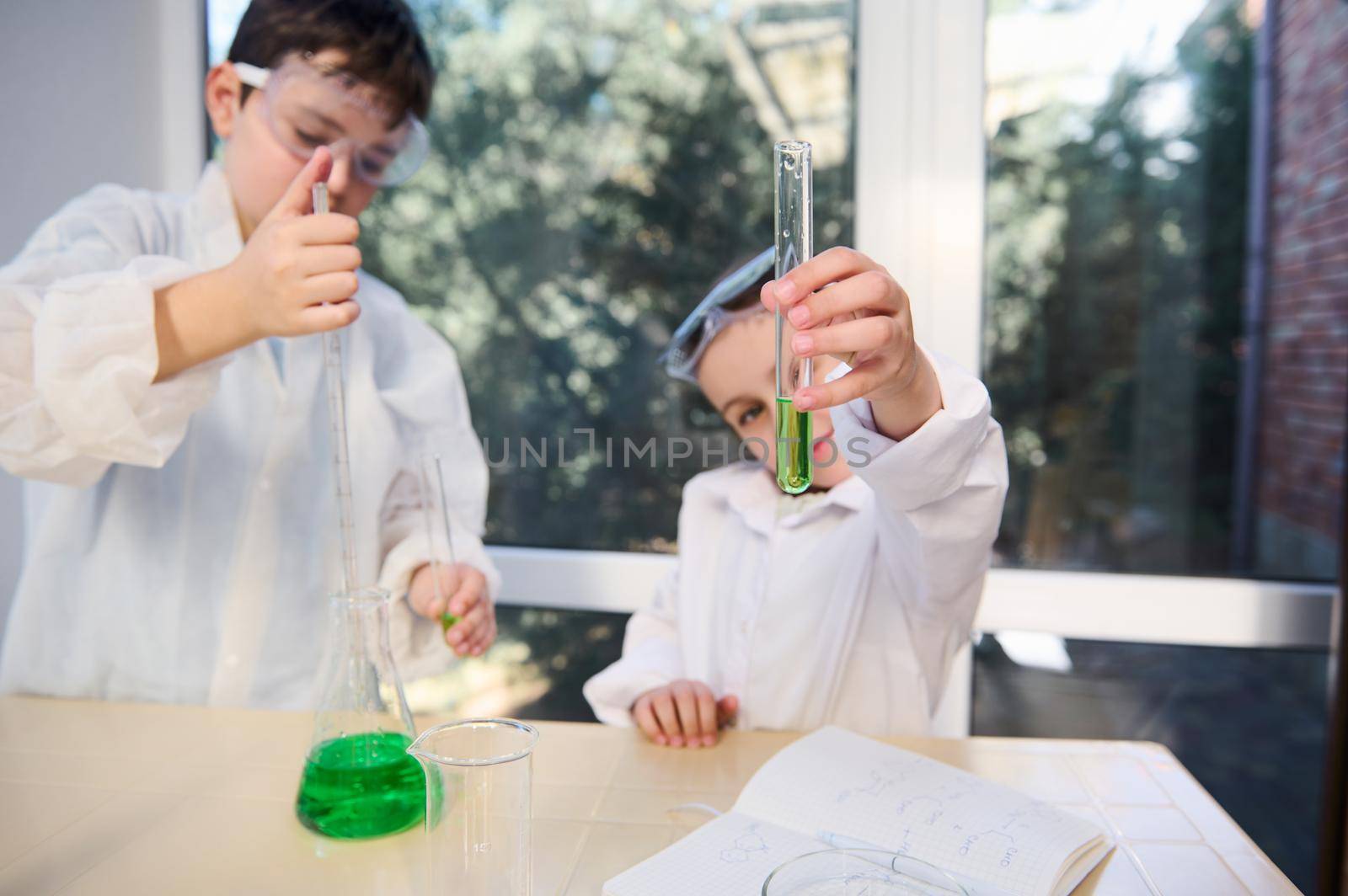 Selective focus on a test tube with green chemical solution in the hands of blurred child girl, watching the going on chemical reaction. Kids learning Chemistry. School laboratory. Knowledge Education