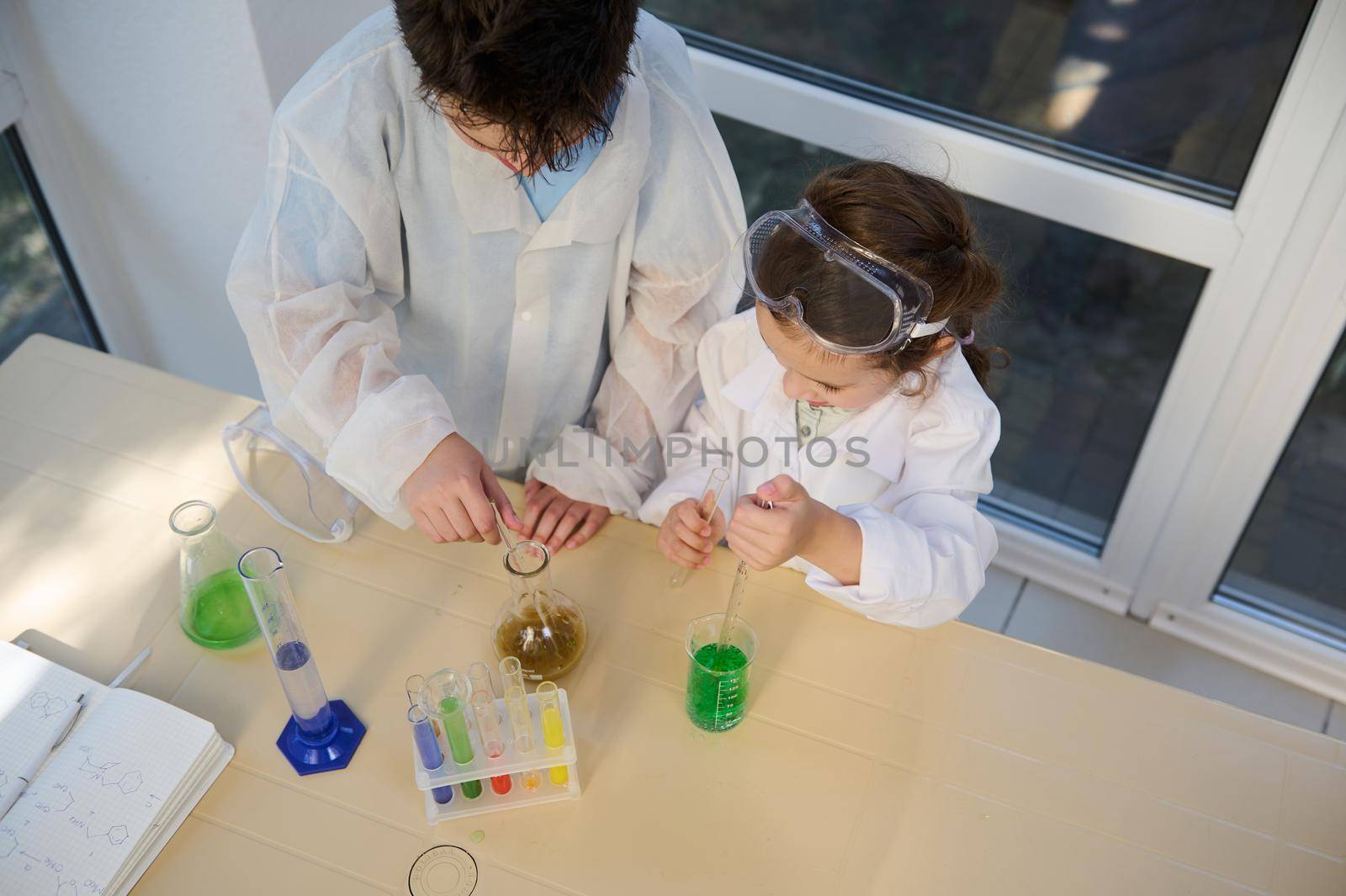 Top view of smart kids pupils, chemist scientist in white lab coats, holding labware with a taking place chemical reaction between chemicals and reagents, conducting experiments in chemistry classroom