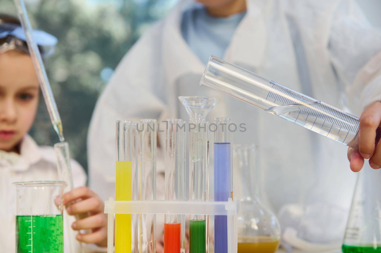 Selective focus on measuring cylinder in the hands of school child, pouring water into test tubes with colorful chemical liquids, during chemistry lesson. Back to school in new academic year semester
