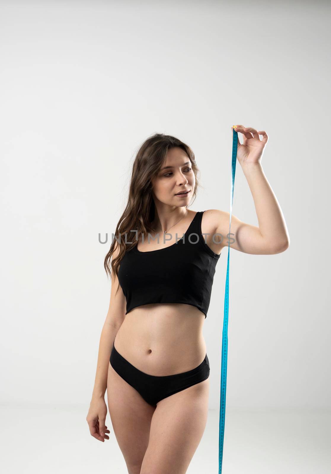 Confused brunette female with slim body in a black underwear looking on a blue measure tape. Healthy nutrition and weight losing concept. Weight loss concepts. by vovsht