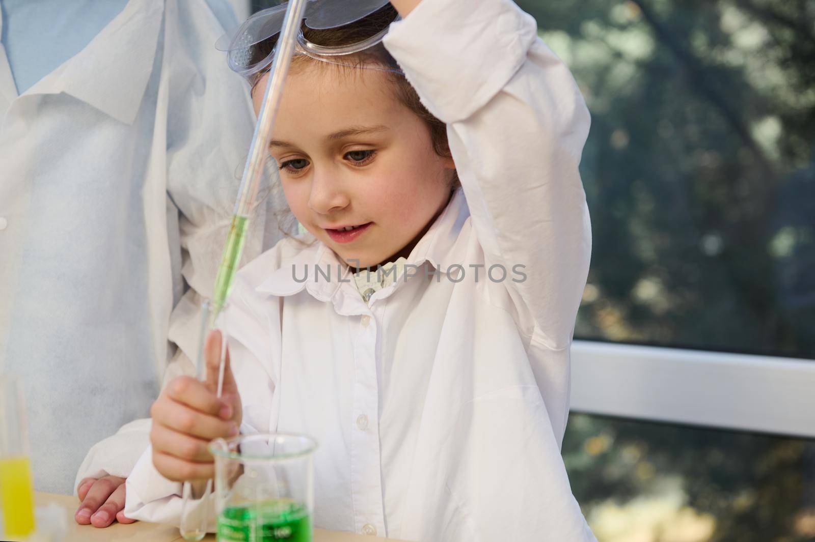 Smart Caucasian schoolgirl, elementary age student wearing white lab coat and goggles, conducts a chemical experiment in school Chemistry laboratory. Education, kids entertainment and development