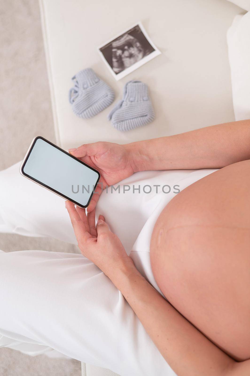 Faceless pregnant woman sits on a white sofa and holds a smartphone. Fetal ultrasonic screening