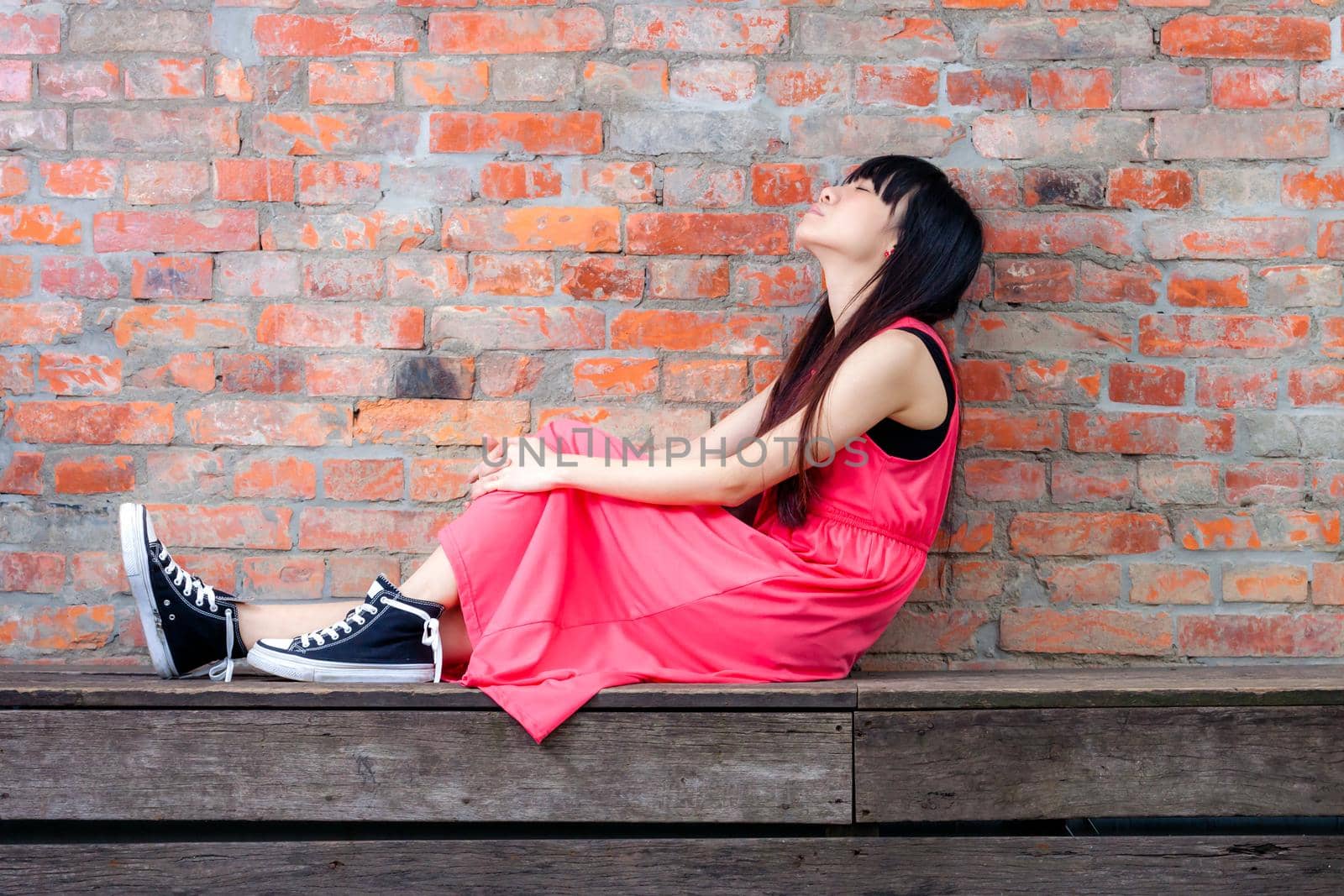 Young Asian woman sitting by red brick wall feeling pensive by imagesbykenny
