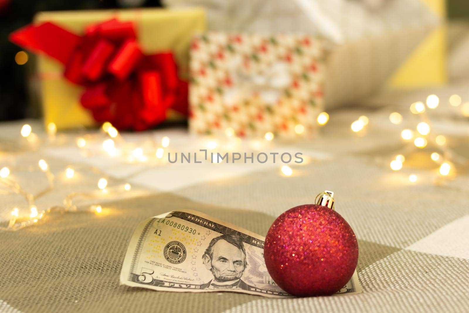 Rolled-up US 5 dollar bill with red shiny christmas decoration, gifts with bright lights and xmas tree on blurred background, festive greeting card, selective focus