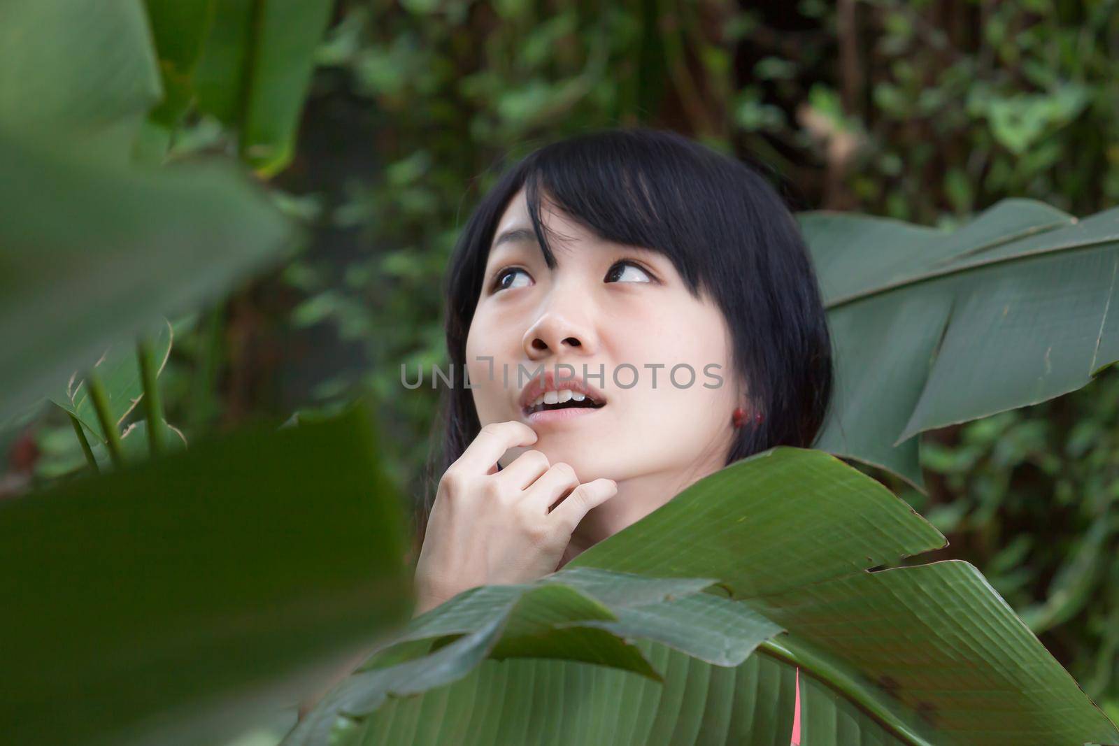 Beautiful Asian girl by green foliage with hand on chin, portrait