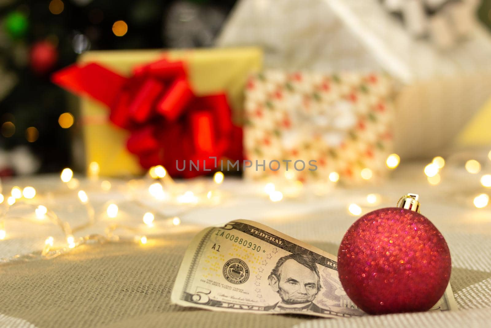 Rolled-up US 5 dollar bill with red shiny christmas decoration, gifts with bright lights and xmas tree on blurred background, festive greeting card, low angle shot with selective focus