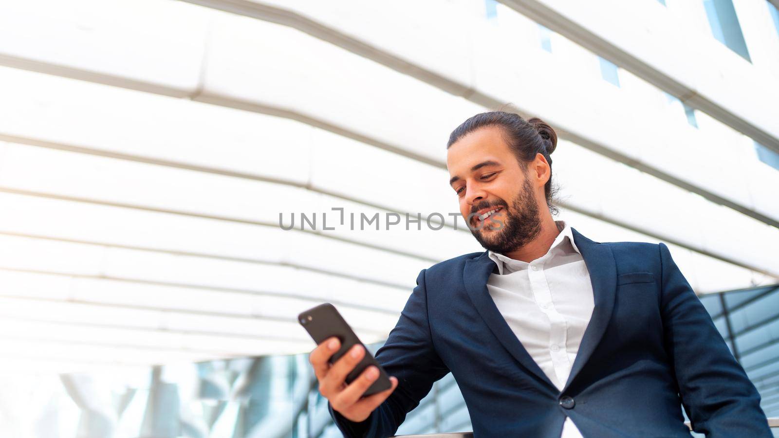Businessman standing outdoor use smartphone looking device screen and smile. Handsome hispanic male business person in business suit holding smartphone in hand. Modern business person low angle view