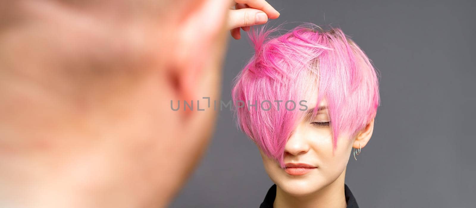 Hairdresser with hands is checking out and fixing the short pink hairstyle of the young white woman in a hair salon. by okskukuruza