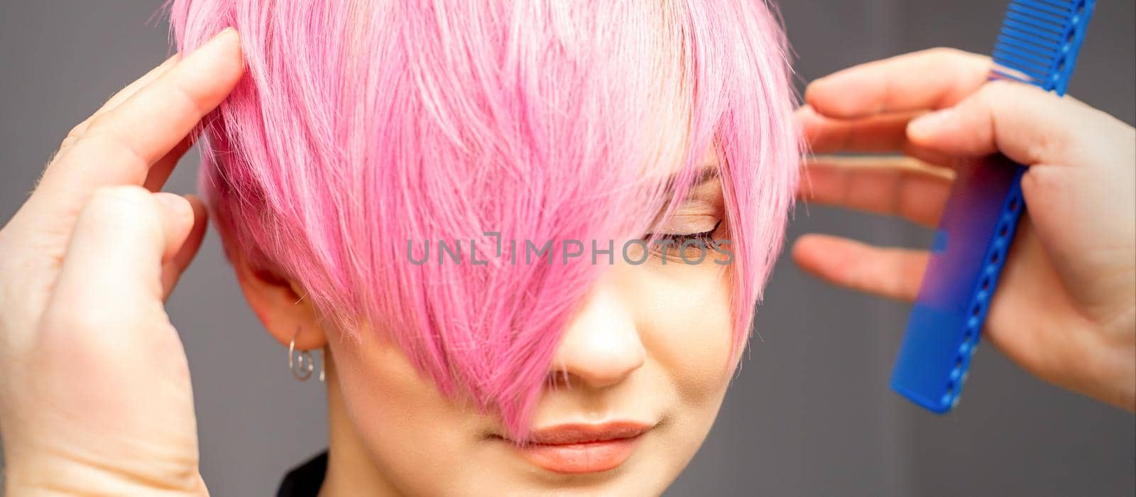 Hairdresser with hands and comb is checking out and fixing the short pink hairstyle of the young white woman in a hair salon. by okskukuruza