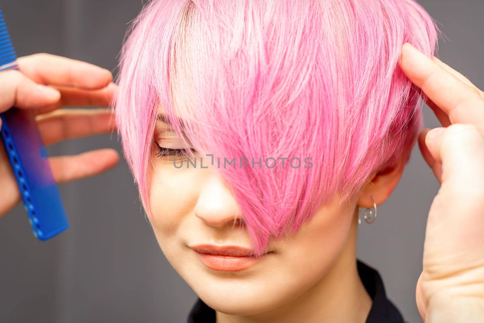 Hairdresser with hands and comb is checking out and fixing the short pink hairstyle of the young white woman in a hair salon