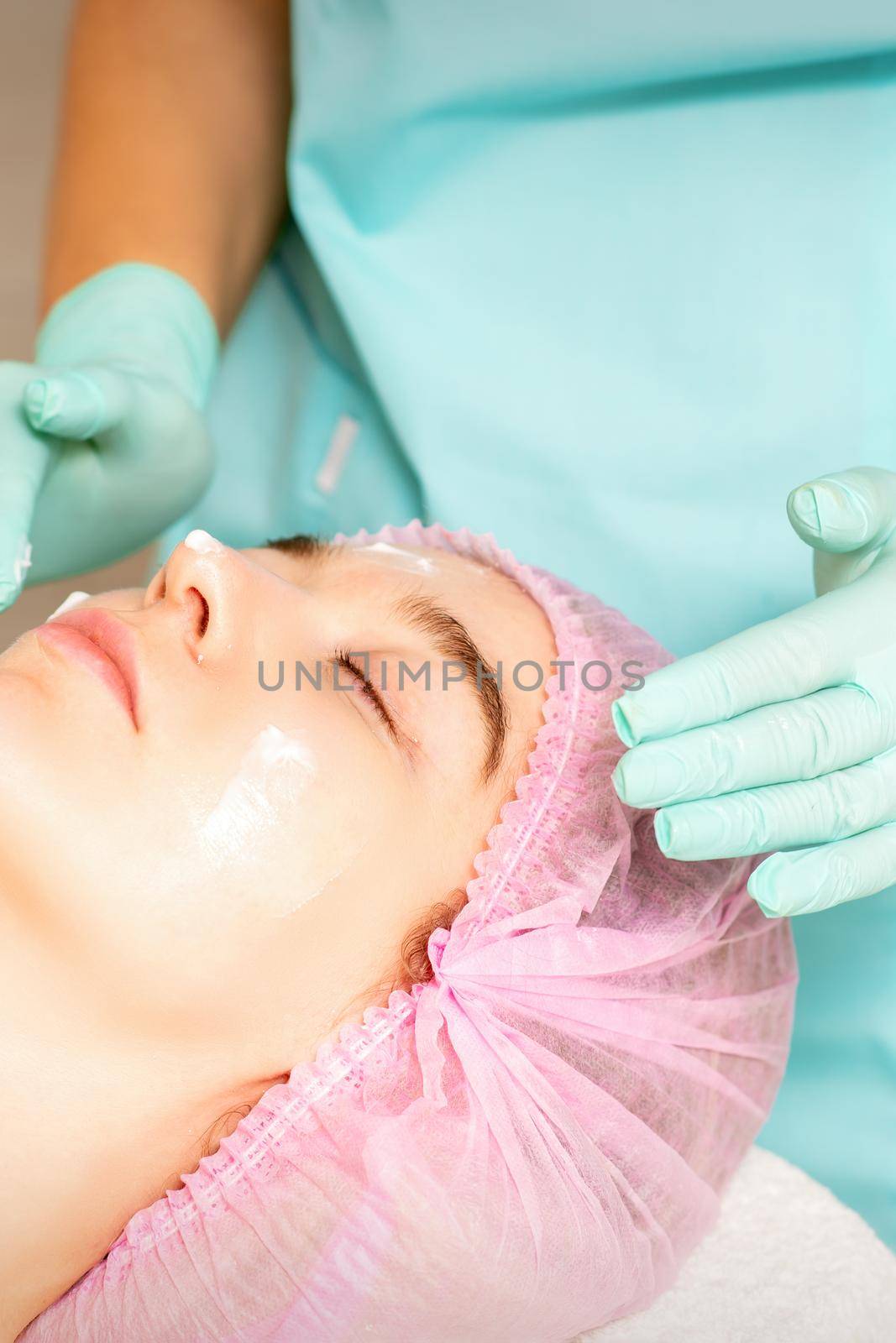 Cosmetologist with gloved hands applies a moisturizing mask with peeling cream on the female face. Facial cosmetology treatment. Procedures for facial care. by okskukuruza