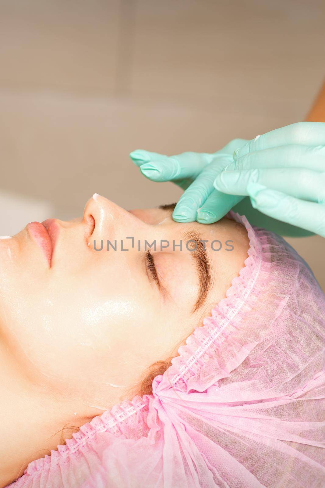 Cosmetologist with gloved hands applies a moisturizing mask with peeling cream on the female face. Facial cosmetology treatment. Procedures for facial care