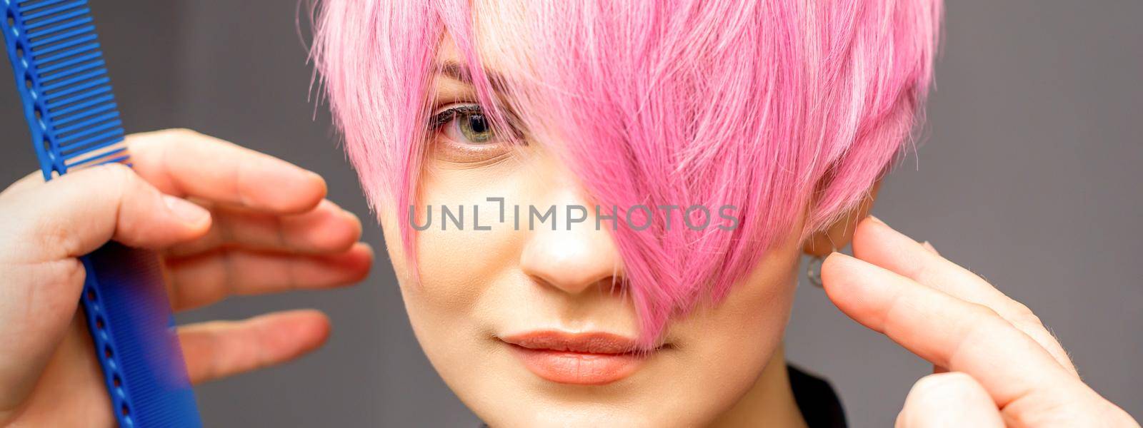 Hairdresser with hands and comb is checking out and fixing the short pink hairstyle of the young white woman in a hair salon. by okskukuruza