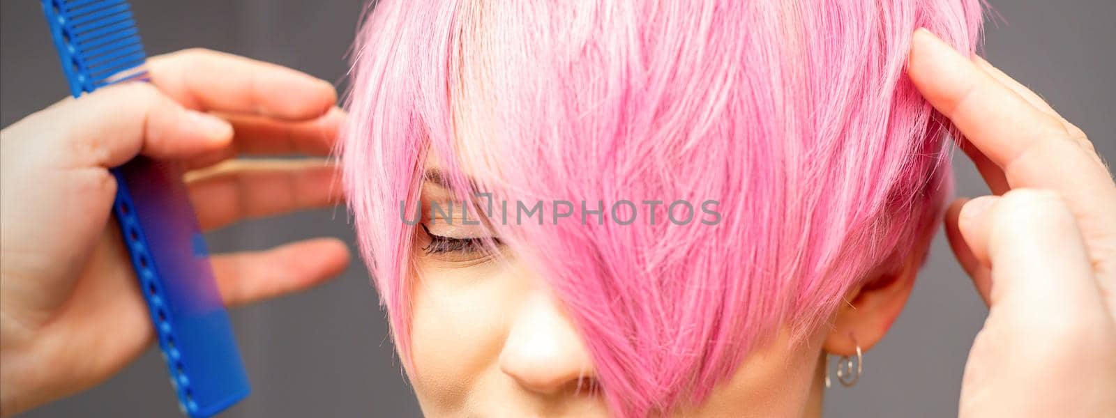 Hairdresser with hands and comb is checking out and fixing the short pink hairstyle of the young white woman in a hair salon