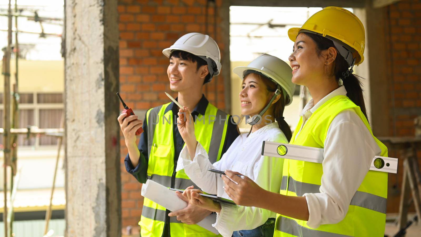 Smiling female investor and engineer managers wearing safety helmet working, checking plan together at construction site.