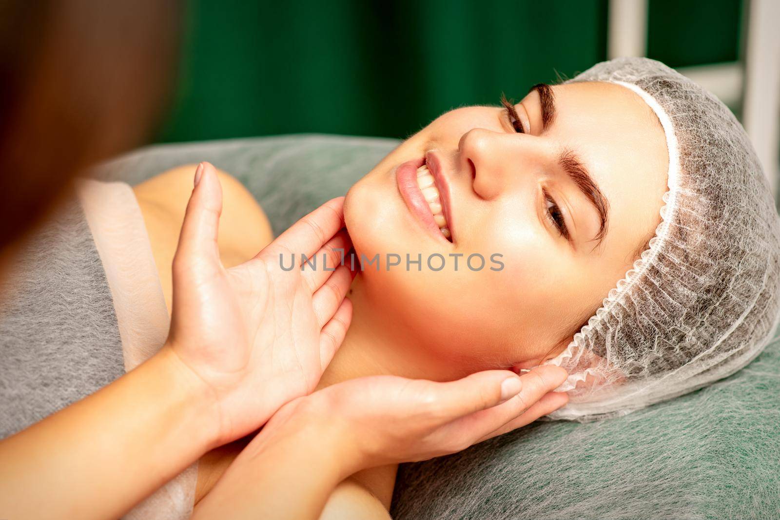 Plastic surgery and beauty concept. Professional cosmetician examining and touching the face of the beautiful young caucasian smiling woman in a clinic of esthetic cosmetology. by okskukuruza