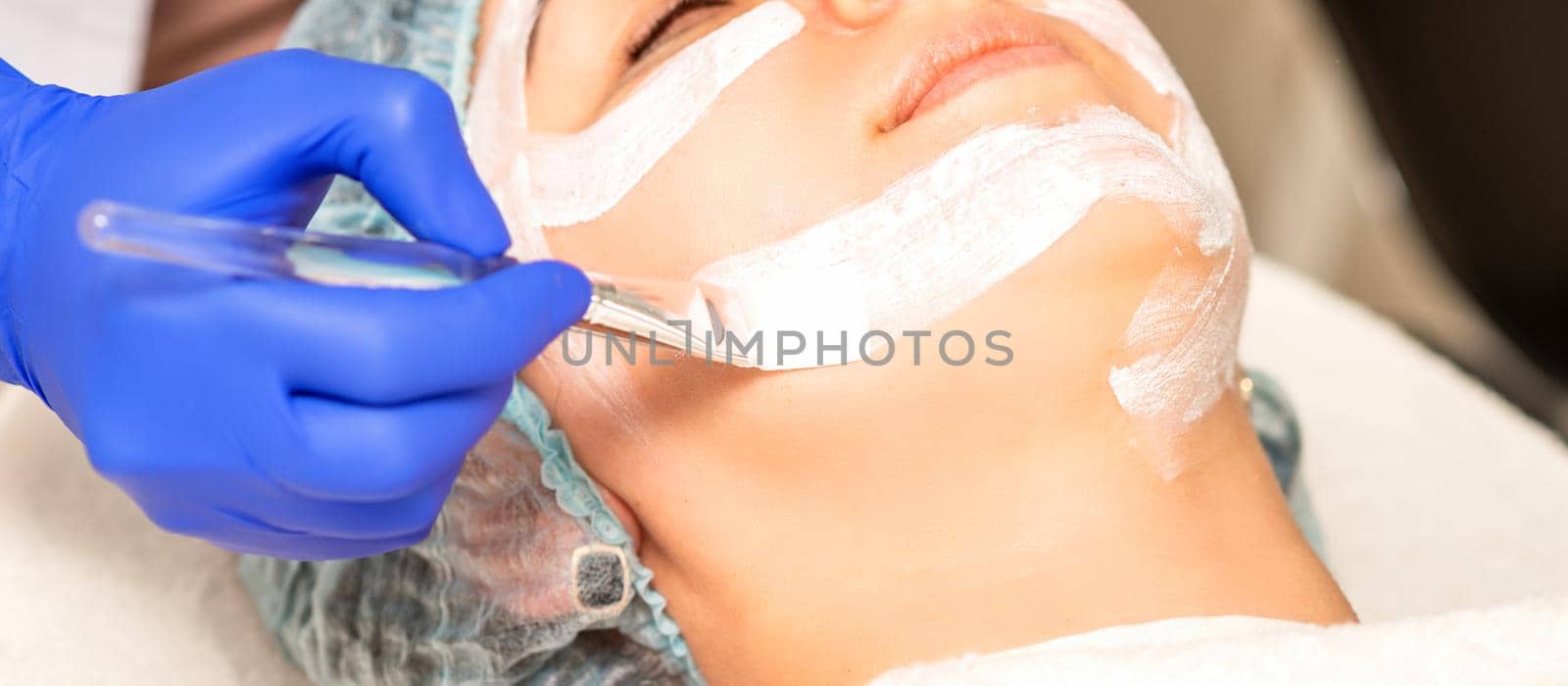 The beautician with brush applies a photochemical and glycolic peeling face mask to the female patient face in the beauty clinic. by okskukuruza