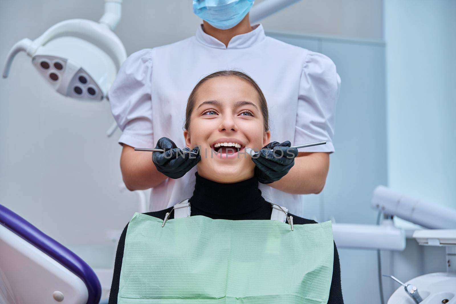 Portrait of young teenage girl in dental chair with hands of doctor with tools. Female teenager smiling with teeth looking at camera in dentist office. Adolescence hygiene treatment dental health care