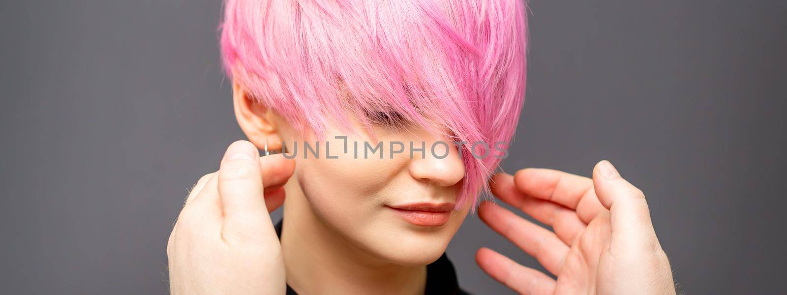 Hairdresser with hands is checking out and fixing the short pink hairstyle of the young white woman in a hair salon