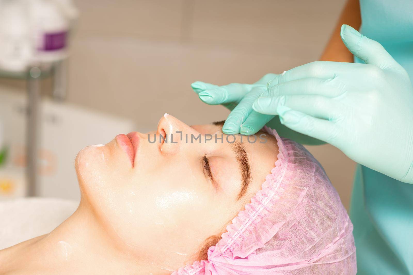 Cosmetologist with gloved hands applies a moisturizing mask with peeling cream on the female face. Facial cosmetology treatment. Procedures for facial care