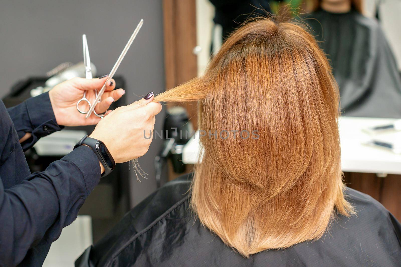 Red-haired woman sitting a front of the mirror and receiving haircut her red long hair by a female hairdresser in a hair salon, back view