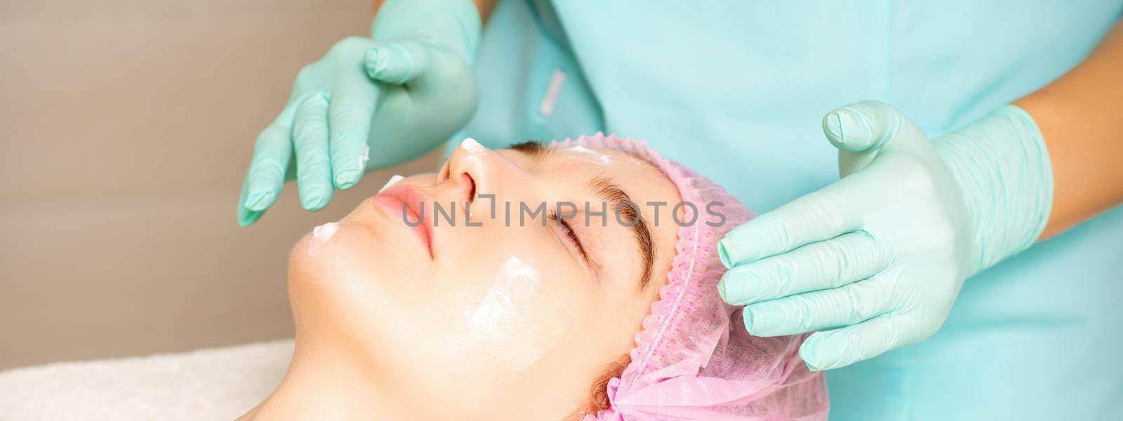 Cosmetologist with gloved hands applies a moisturizing mask with peeling cream on the female face. Facial cosmetology treatment. Procedures for facial care