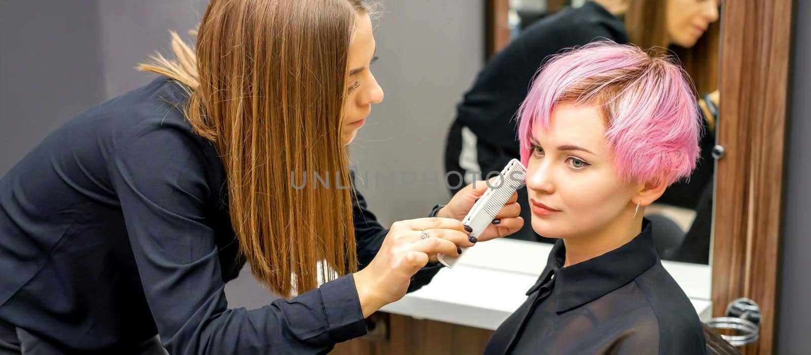 Hairdresser with comb is checking out and fixing the short pink hairstyle of the young white woman in a hair salon