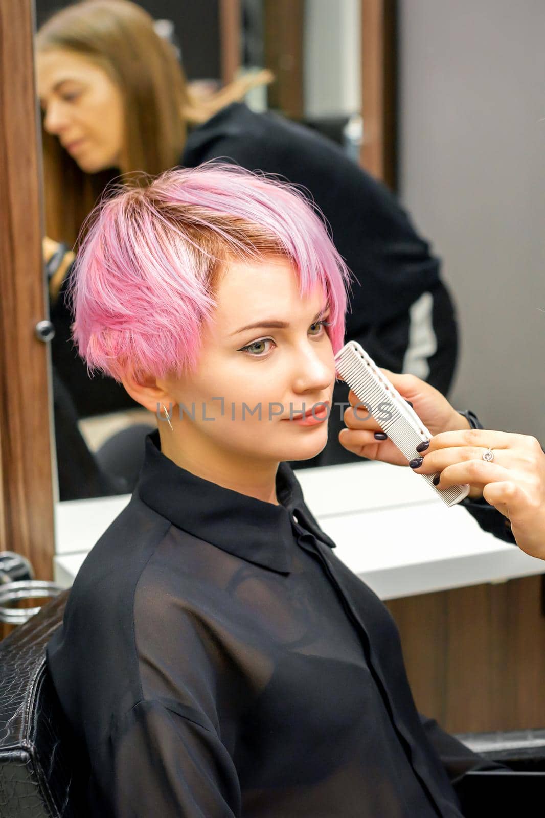 Hairdresser with comb is checking out and fixing the short pink hairstyle of the young white woman in a hair salon