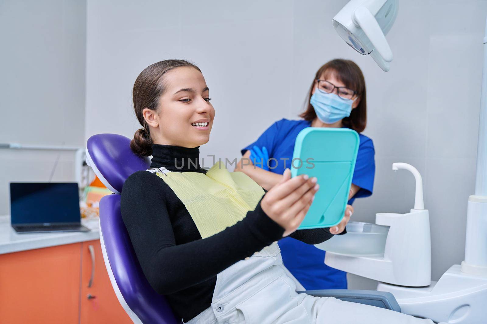 Happy young teenage female with mirror sitting in dentist chair smiling looking at healthy teeth, with dentist doctor in office. Dentistry, hygiene, treatment, dental health care concept