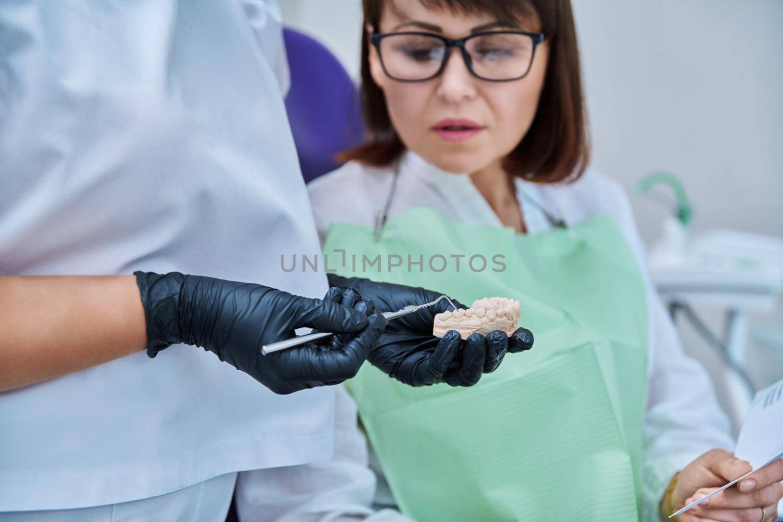 Close-up of plaster model of jaw in hands of doctor, female patient looking and listening to dentist consultation. Dental treatment, prosthetics, implantation, health teeth care concept