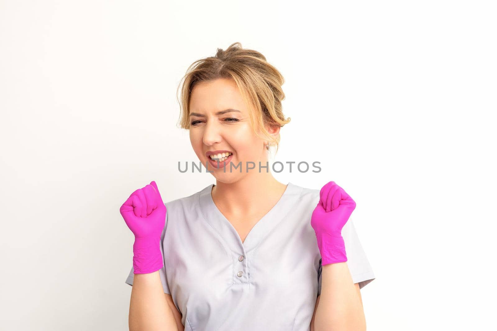 Happy caucasian woman doctor wearing pink gloves celebrates and raising fists on white background. by okskukuruza