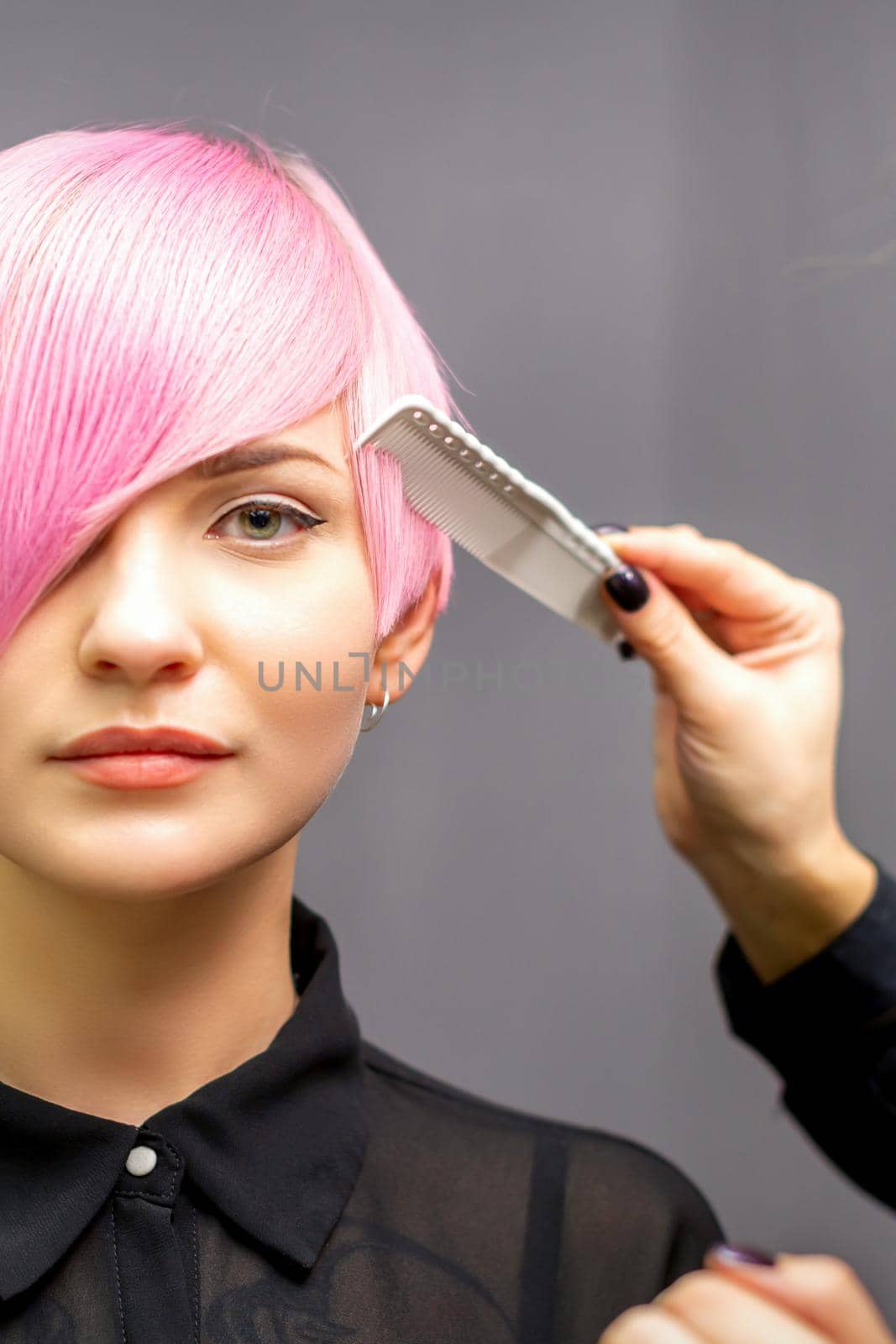 The female hairdresser is combing the dyed pink short hair of the young woman against the dark wall