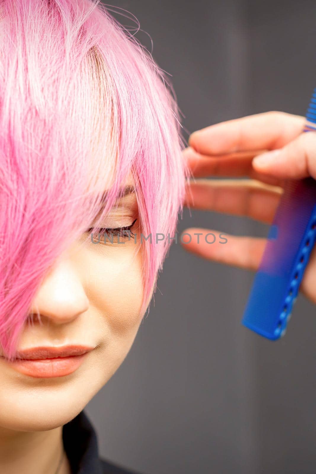 Hairdresser with hands and comb is checking out and fixing the short pink hairstyle of the young white woman in a hair salon