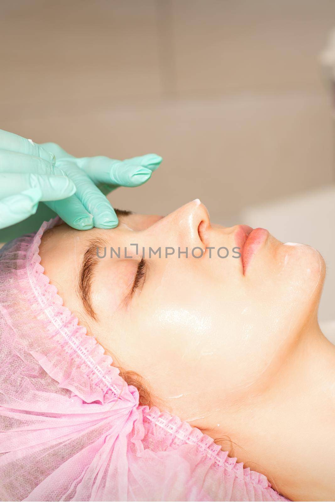 Cosmetologist with gloved hands applies a moisturizing mask with peeling cream on the female face. Facial cosmetology treatment. Procedures for facial care