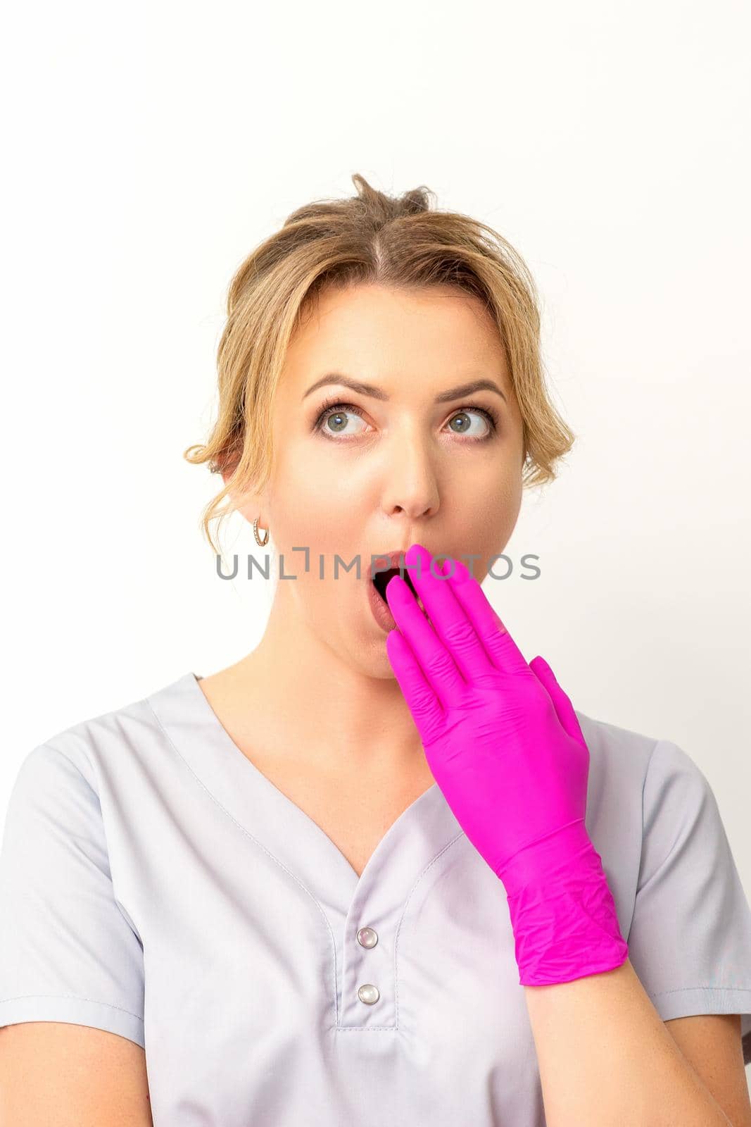 Portrait of a young female caucasian doctor or nurse is shocked covering her mouth with her pink gloved hands against a white background