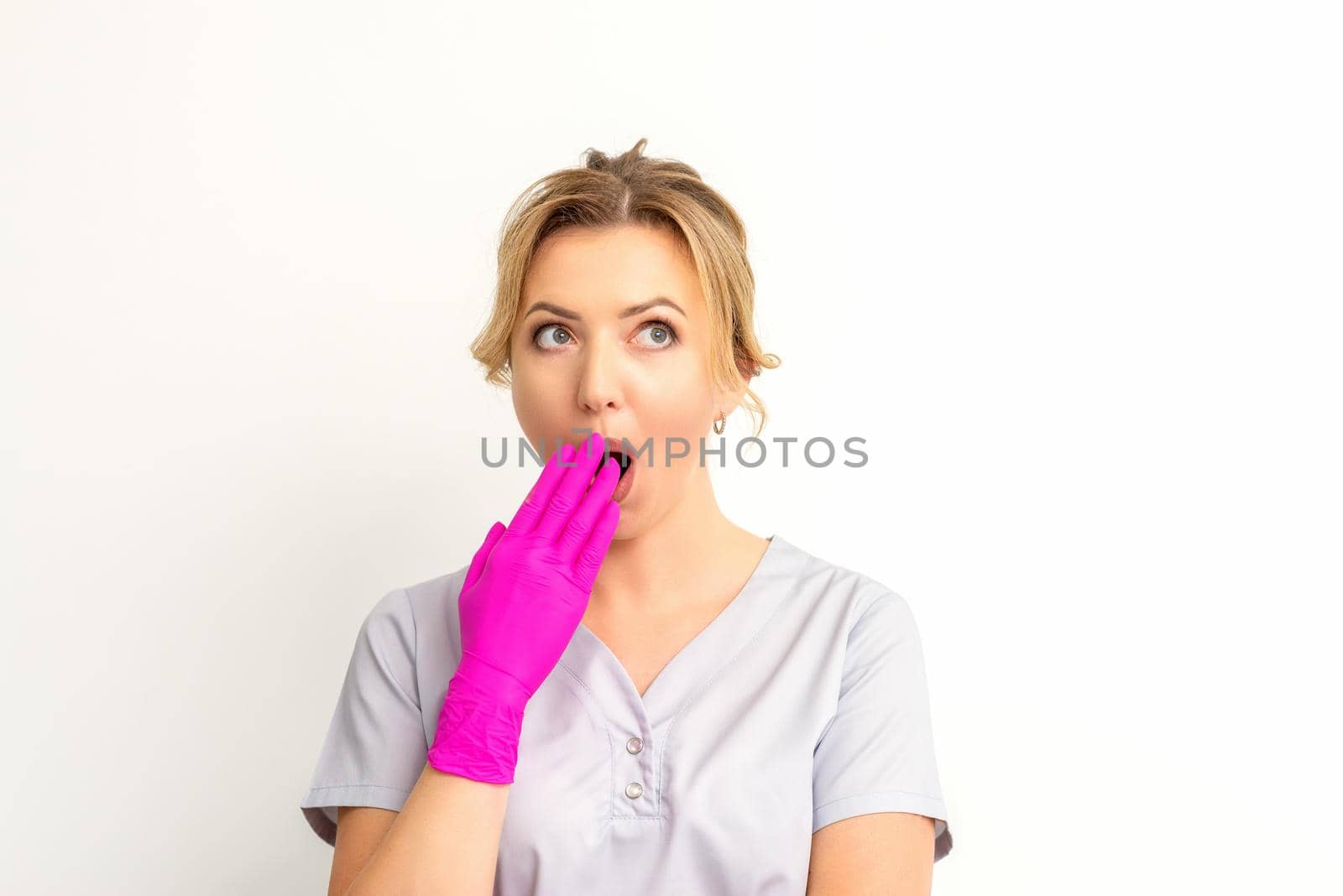 Portrait of a young female caucasian doctor or nurse is shocked covering her mouth with her pink gloved hands against a white background