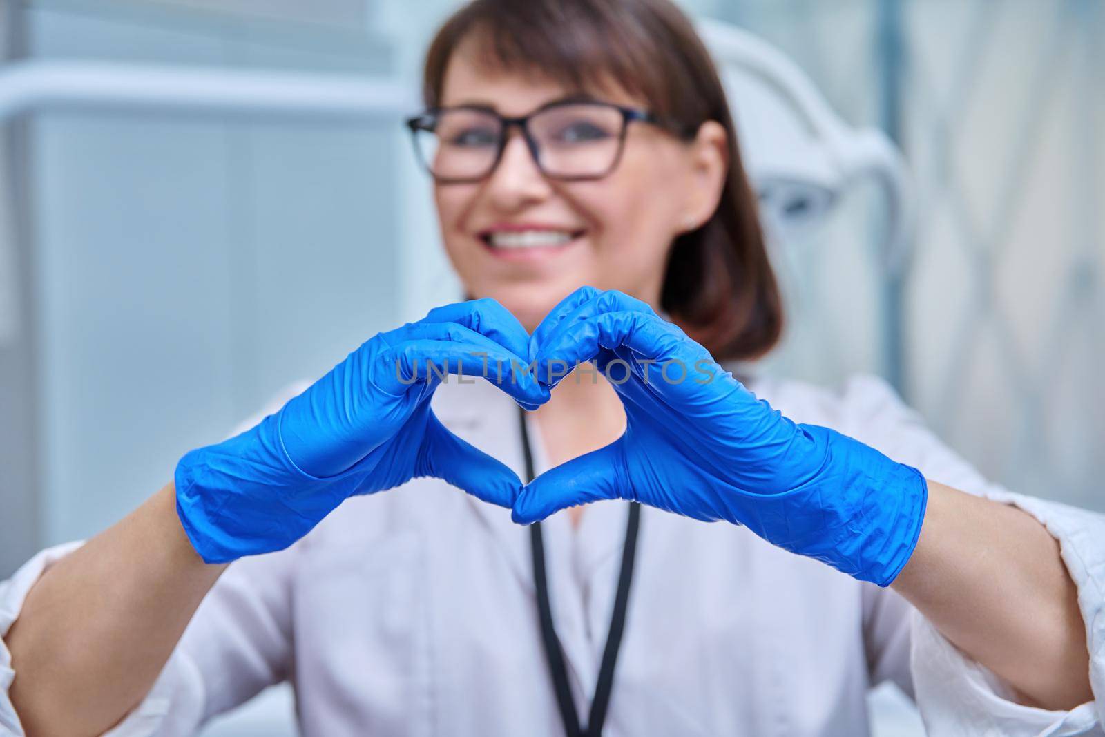Close-up of heart sign symbol shown by nurse doctor dentist in blue protective gloves, dentistry dental office background. Care, love, safety, symbolic gesture of a medical worker to patients