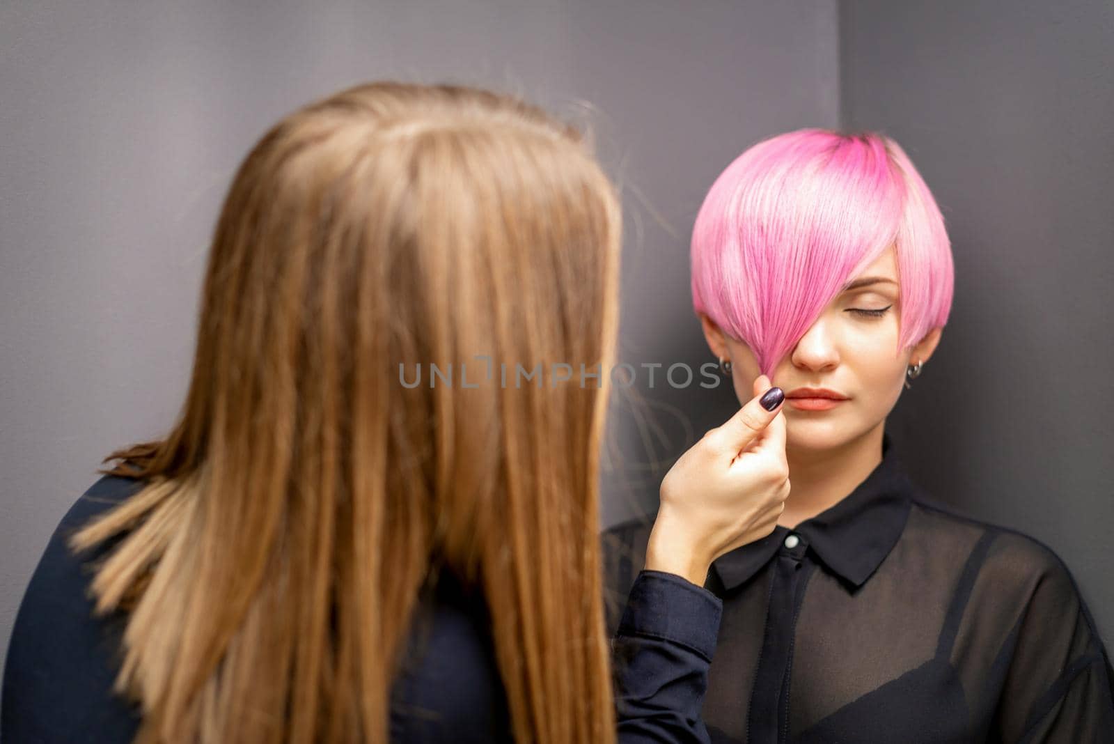 Hairdresser with hands is checking out and fixing the short pink hairstyle of the young white woman in a hair salon