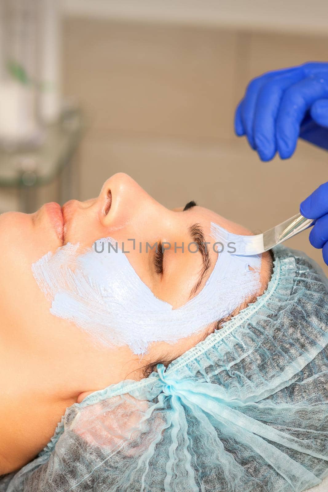 The beautician with brush applies a photochemical and glycolic peeling face mask to the female patient face in the beauty clinic