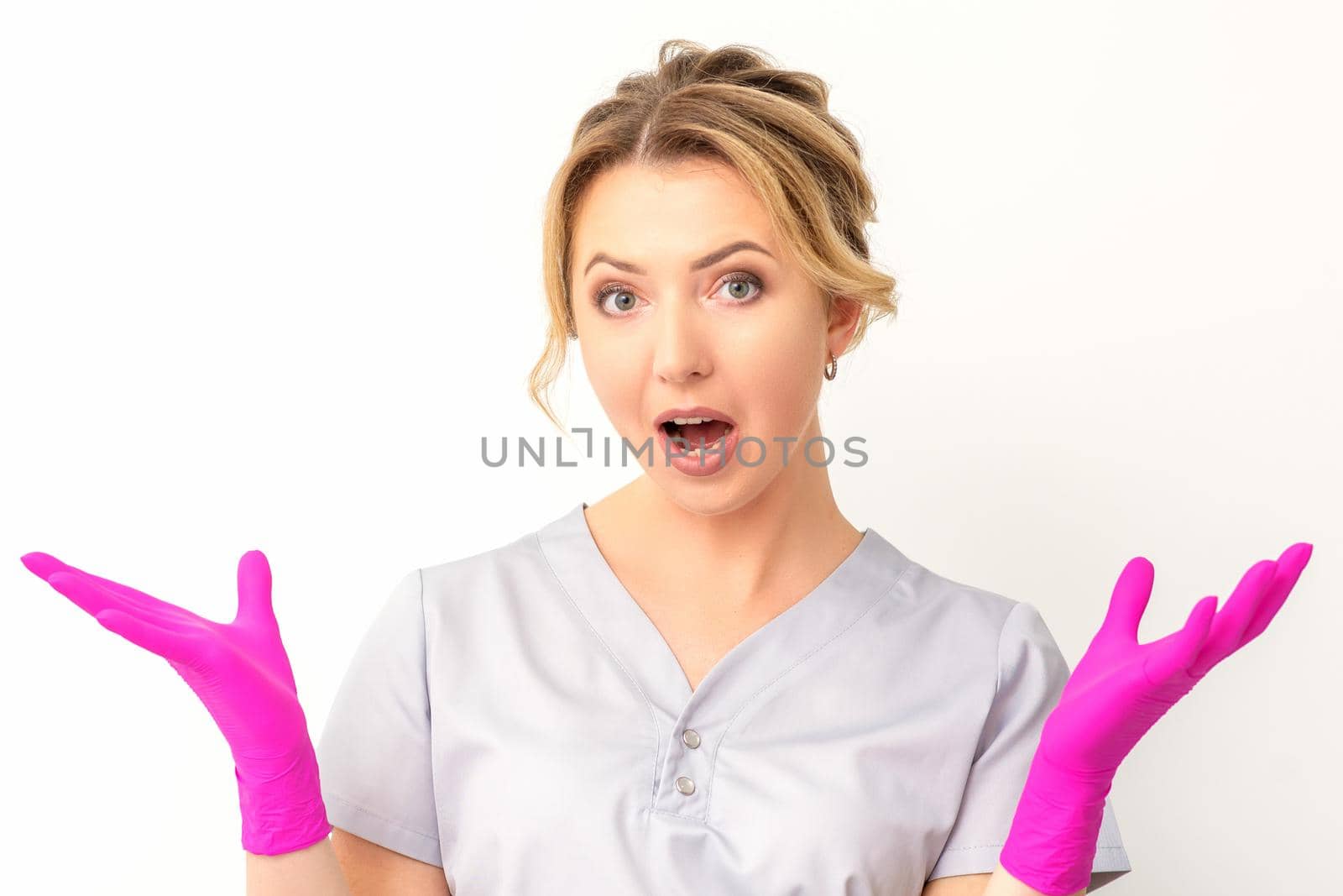 Body language. Young surprised Caucasian woman doctor wearing gloves gesturing with her hands, spread his arms having shocked expression opening her mouth against the white background