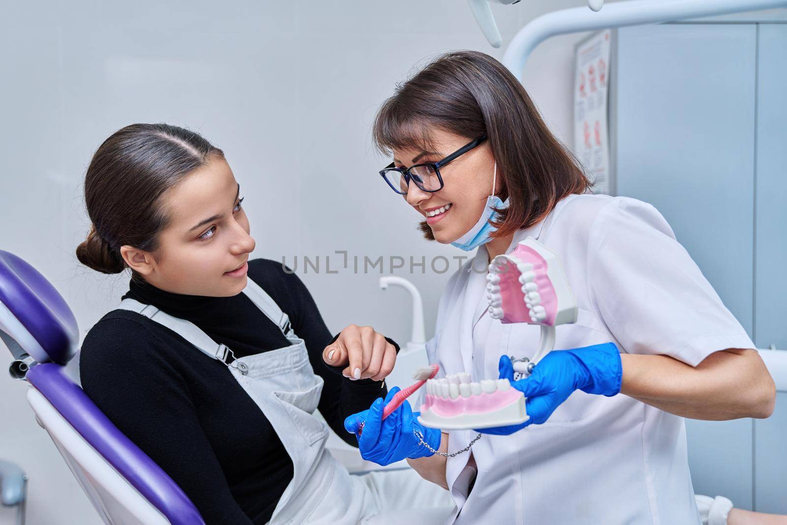 Teenage female sitting in dental chair, doctor with jaw model and toothbrush by VH-studio