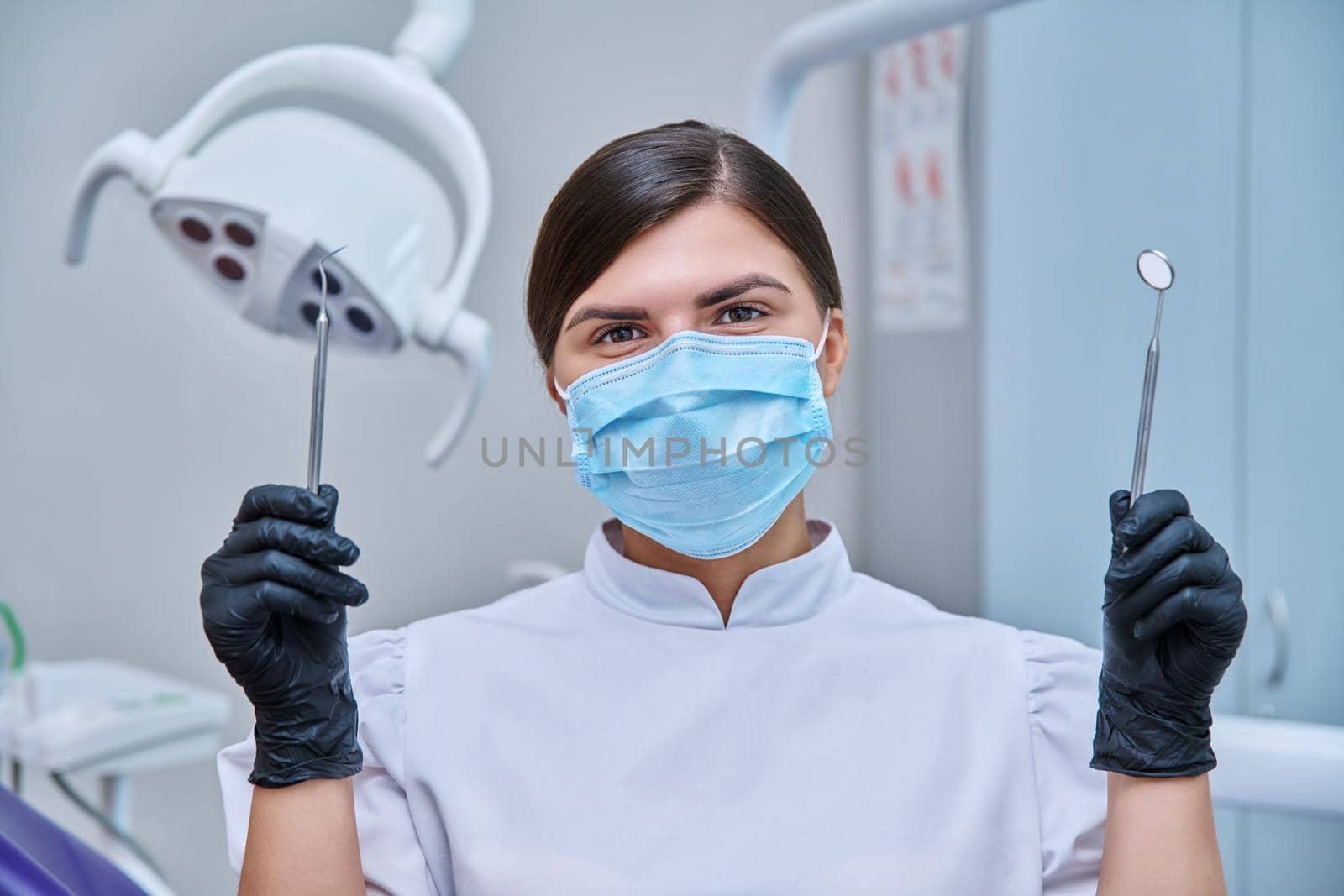 Headshot portrait of female dentist wearing facial mask with instruments for examining teeth. Doctor looking at camera, posing in dental office. Dentistry, medicine, treatment, dental health care