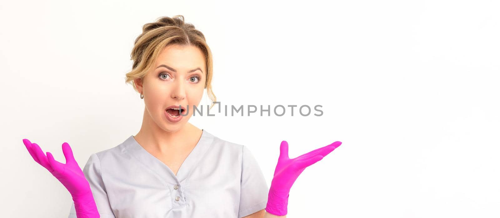 Body language. Young surprised Caucasian woman doctor wearing gloves gesturing with her hands, spread his arms having shocked expression opening her mouth against the white background