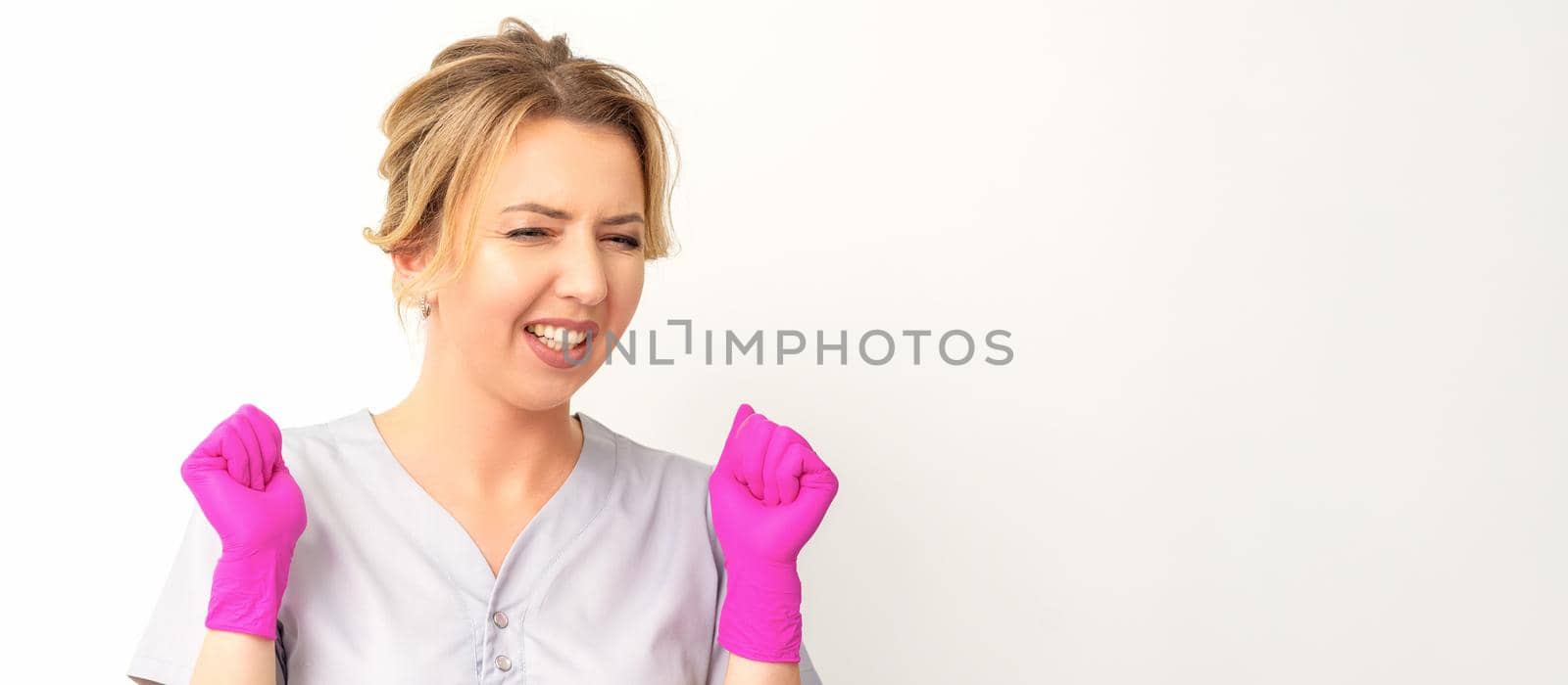 Happy caucasian woman doctor wearing pink gloves celebrates and raising fists on white background