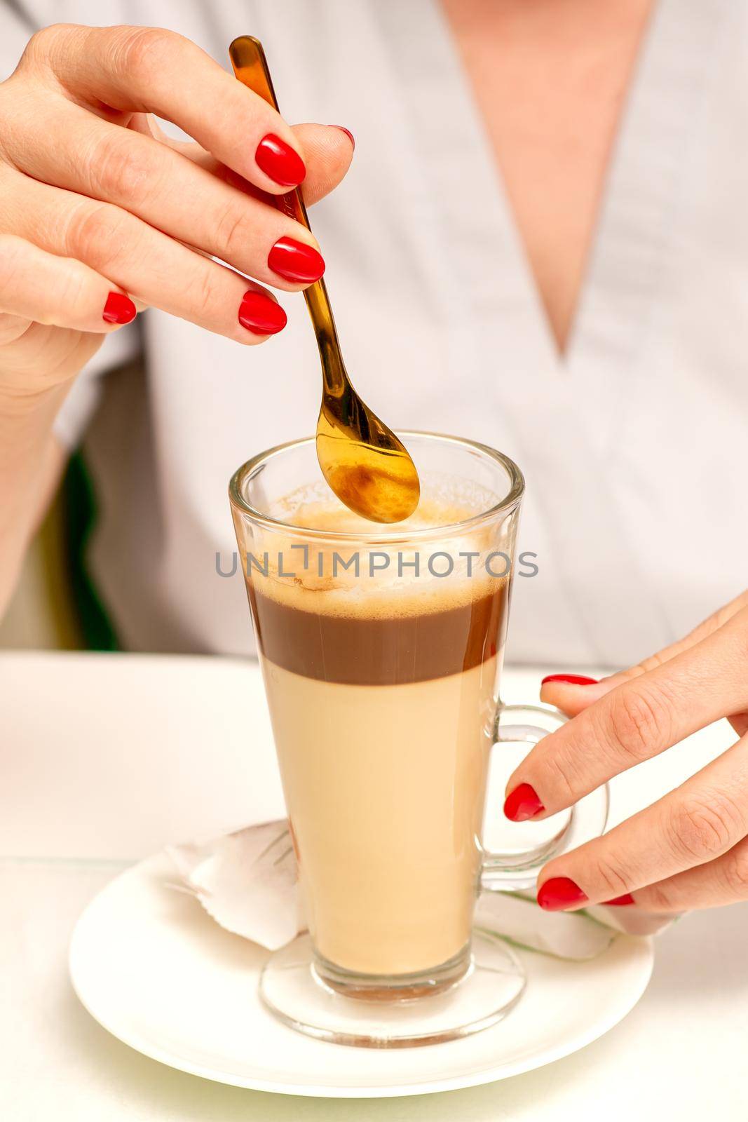 Woman with the latte. Glass mug of latte coffee on the white saucer with female hands holding teaspoon on the table in the cafeteria, close up. by okskukuruza