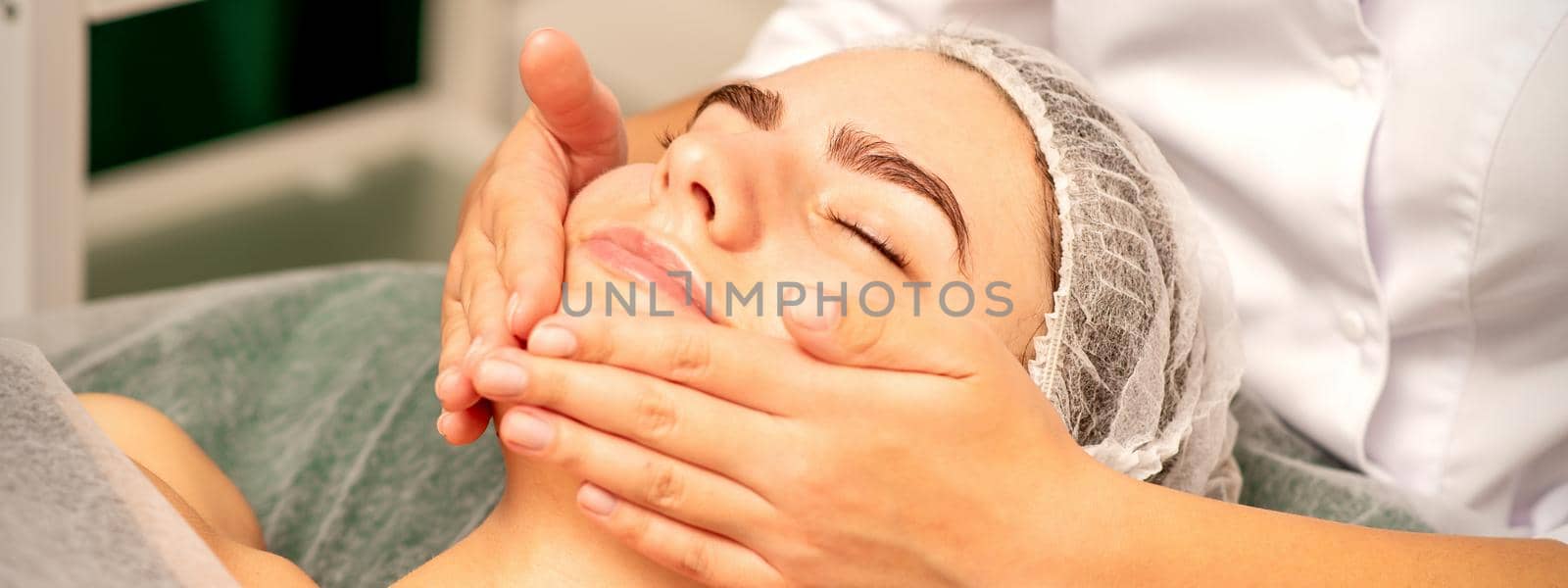 Beautiful caucasian young woman receiving a facial massage with closed eyes in spa salon, close up. Relaxing treatment concept. by okskukuruza