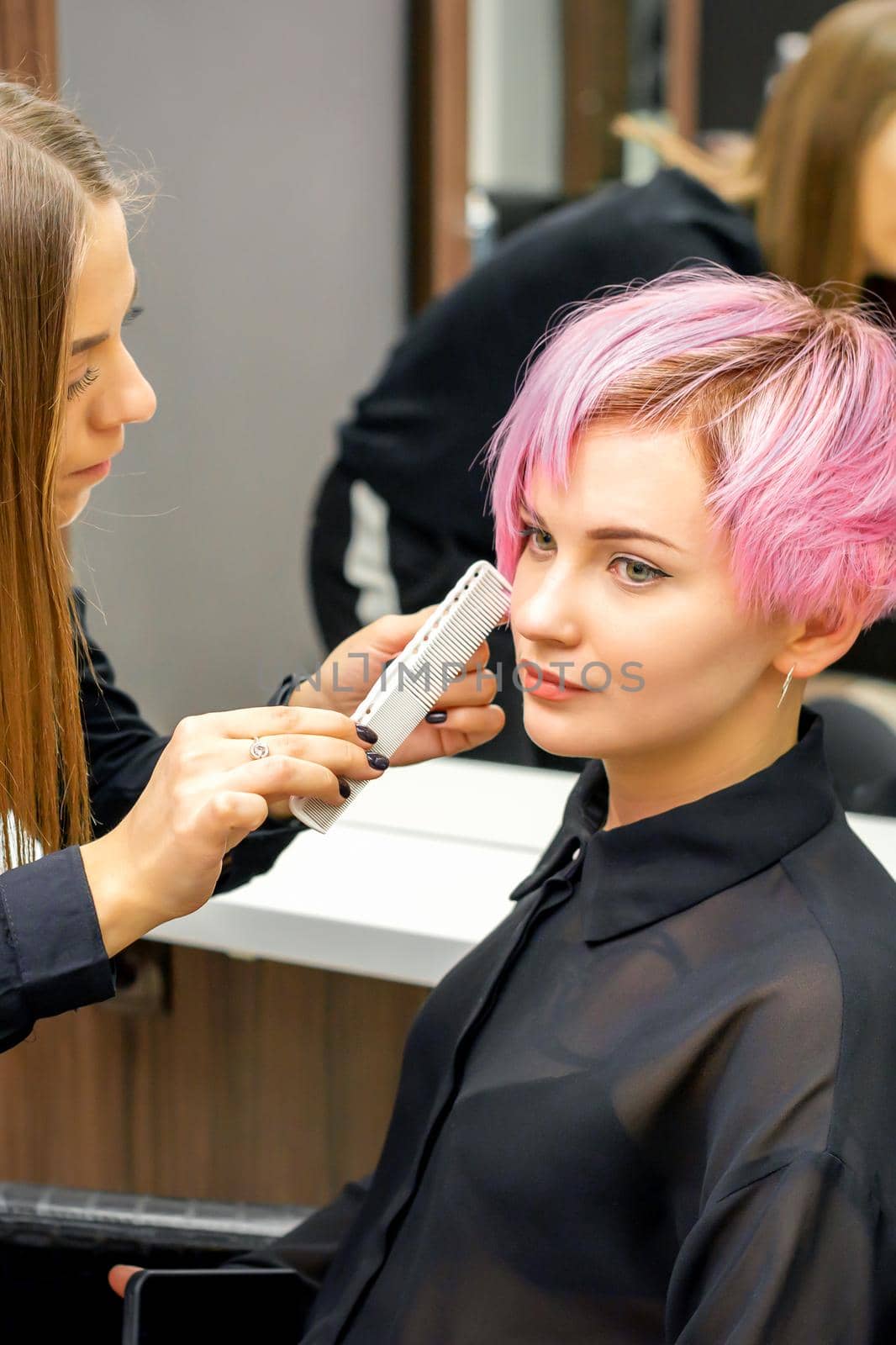 Hairdresser with comb is checking out and fixing the short pink hairstyle of the young white woman in a hair salon. by okskukuruza